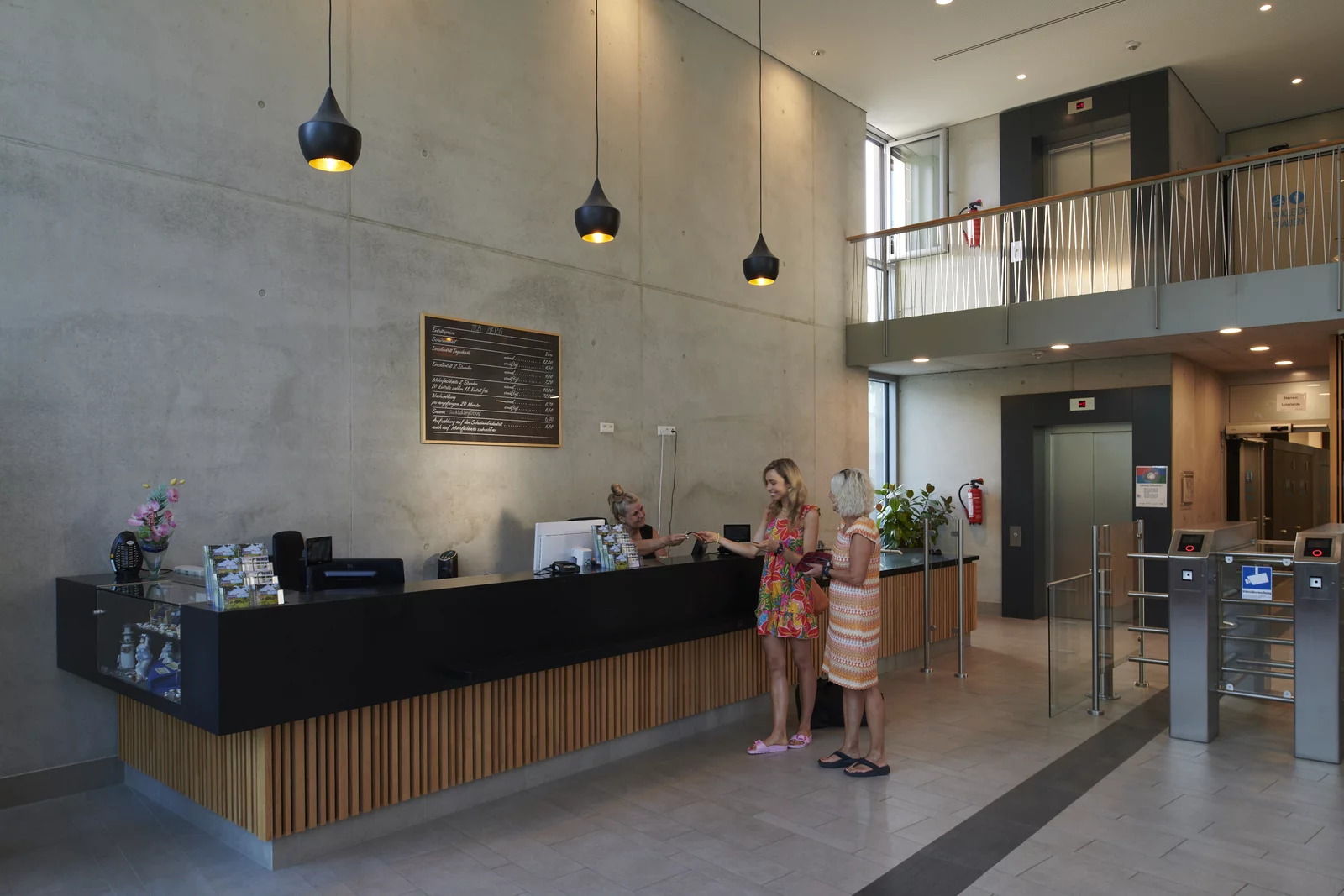 Zwei Frauen kaufen Eintrittstickets an der Kasse im Foyer im Mineralbad Berg. Die Kassiererin reicht einer jüngen Frau das Armband für den Eintritt ins Bad.