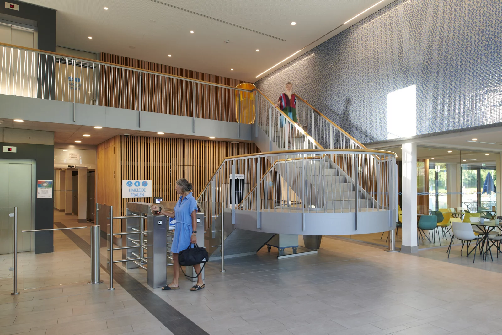 Eine Frau geht durch die Schranke im Foyer ins Bad. Eine weitere Frau schreitet die Treppe, vorbei an der goldenen Badewanne hinunter.