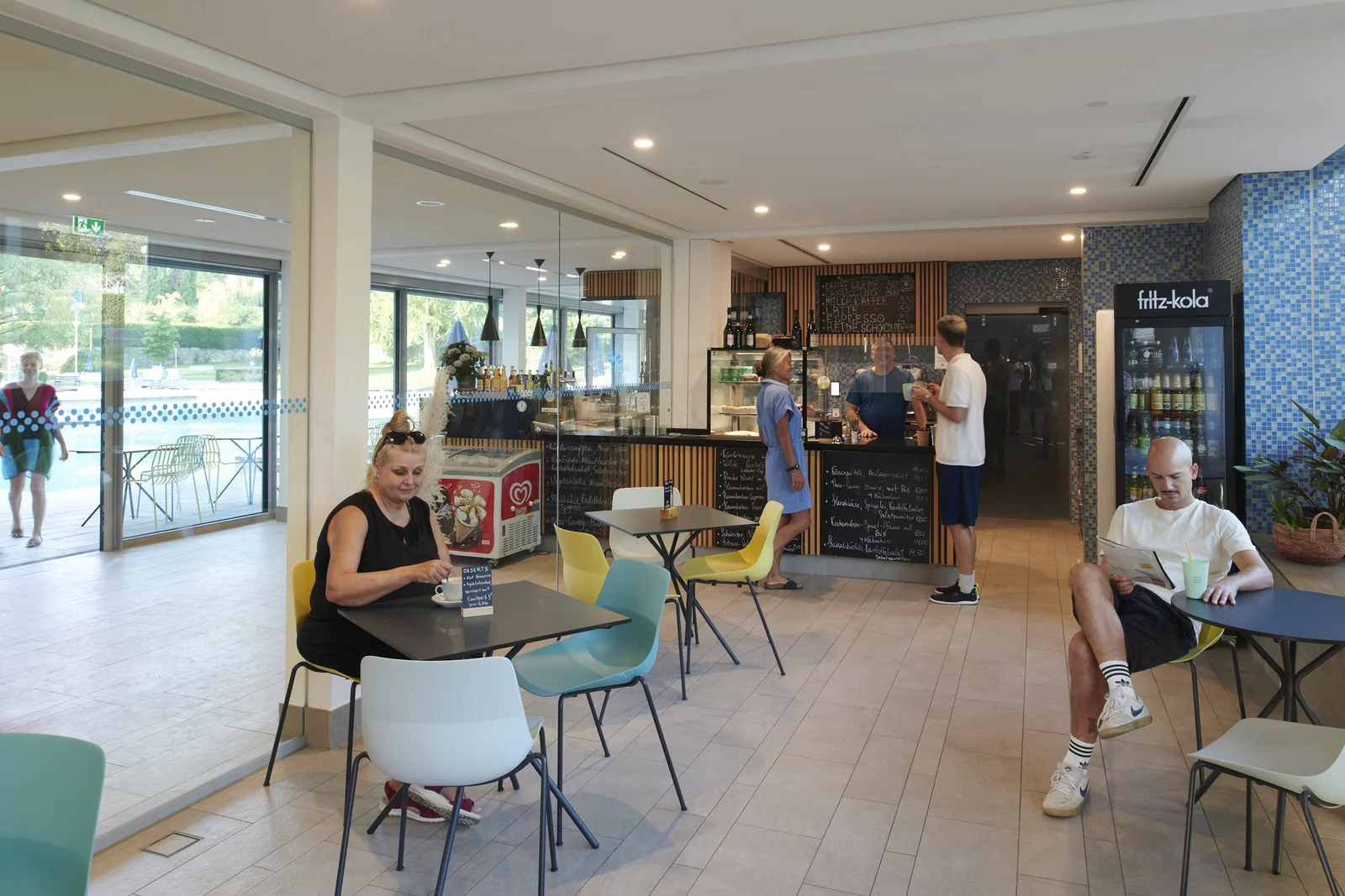 Gemütliches Café im Mineralbad Berg. Eine Frau sitzt am Tisch links im Bild. Rechts blättert ein jünger Mann am Tisch eine Zeitschrift. Im Hintergrund ist eine Theke zu sehen.